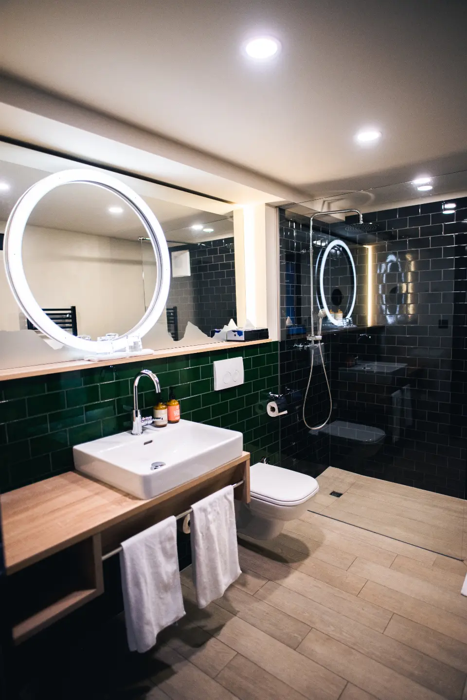 A bathroom with dark green tiles, a round lit mirror and a timber vanity