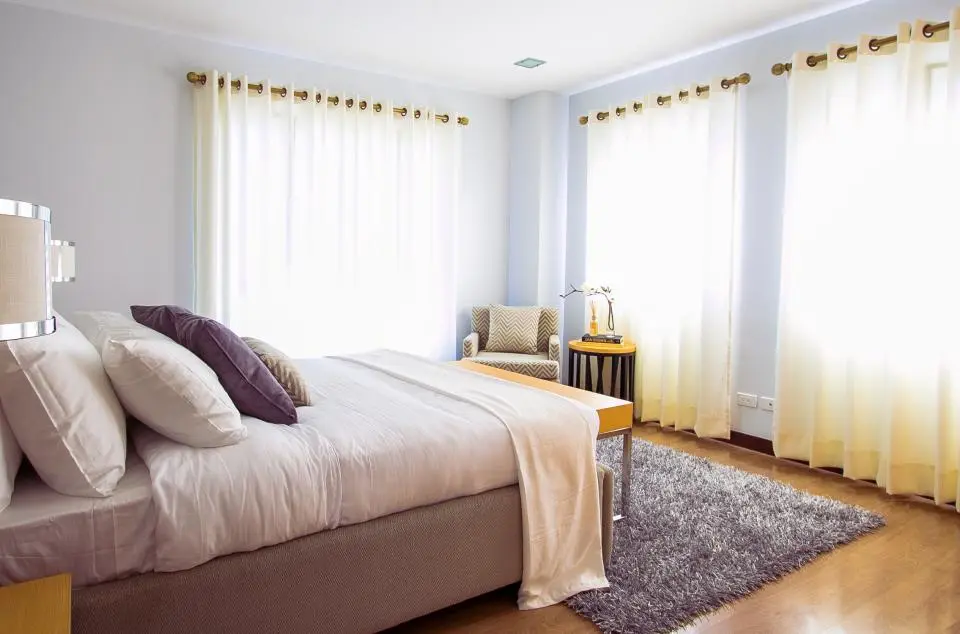 A light bedroom with sheer curtains, a timber floor and plum cushions